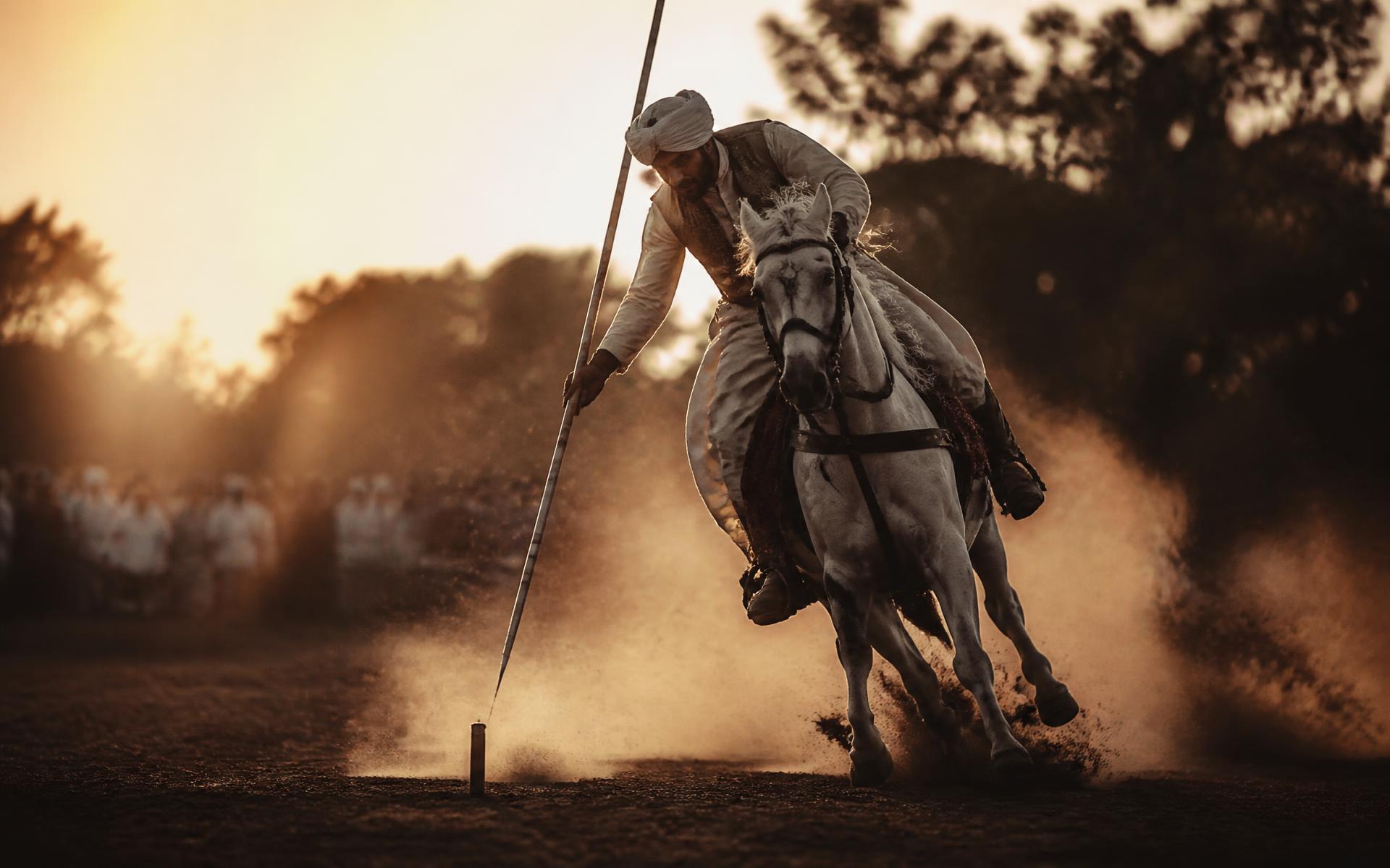 Tent pegging rider galloping with lance lowered toward a ground peg at sunset